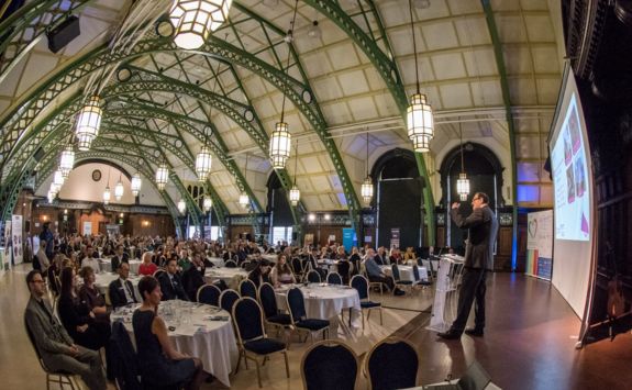 Charles Harvey speaking on stage at the Generosity Festival launch event held at the Discovery Museum. The audience is seated at round tables under an ornate arched ceiling with large hanging lights.